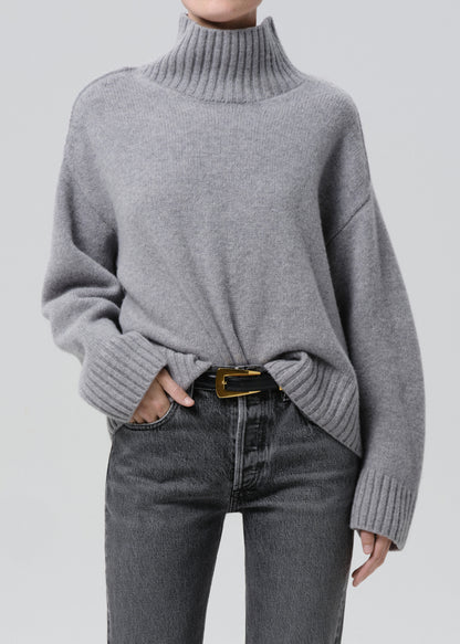 Person wearing a light grey oversized turtleneck sweater with grey straight leg jeans on a grey background
