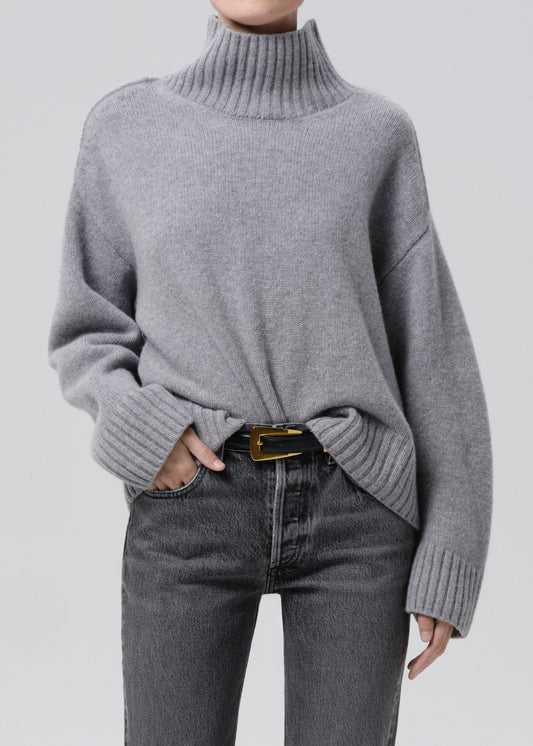 Person wearing a light grey oversized turtleneck sweater with grey straight leg jeans on a grey background