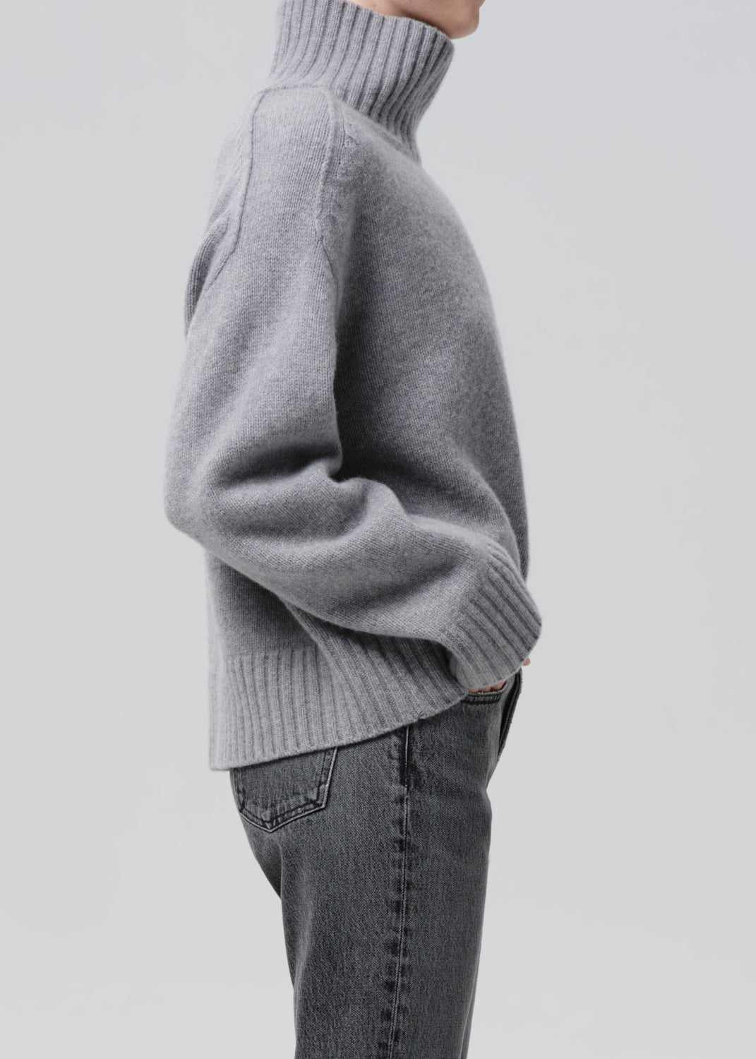 Person wearing a light grey oversized turtleneck sweater with grey straight leg jeans viewed from the side on a grey background