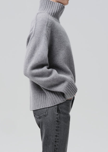 Person wearing a light grey oversized turtleneck sweater with grey straight leg jeans viewed from the side on a grey background