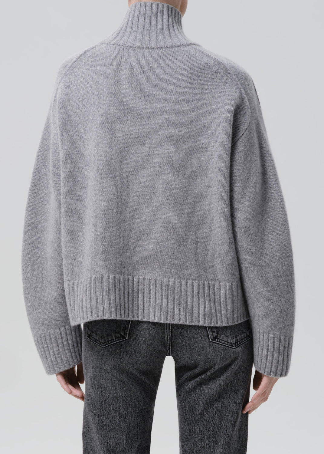 Person wearing a light grey oversized turtleneck sweater with grey straight leg jeans viewed from the back on a grey background