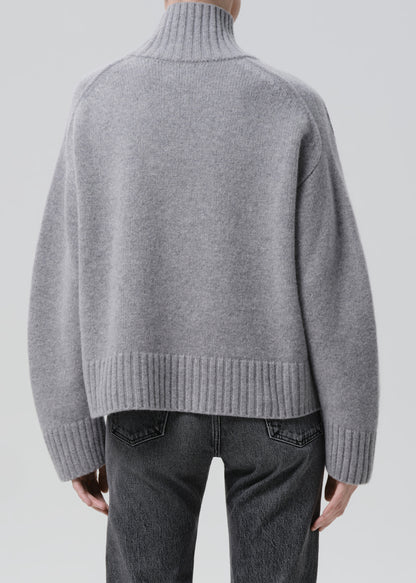 Person wearing a light grey oversized turtleneck sweater with grey straight leg jeans viewed from the back on a grey background