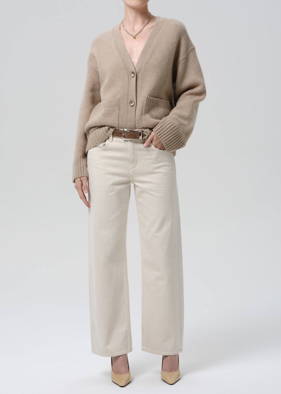 Person wearing a beige cardigan and white pants on a grey background