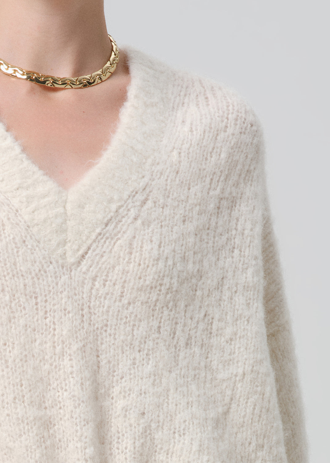 Close-up of a person wearing a white knitted sweater and gold necklace on a grey background