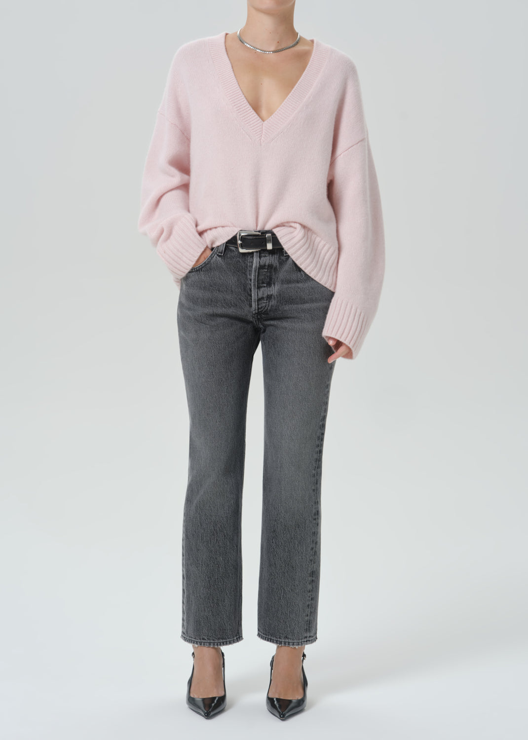 A model wearing a light pink v-neck knit sweater, dark grey cropped jeans, and black pointed-toe heels against a light grey background.