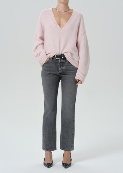 A model wearing a light pink v-neck knit sweater, dark grey cropped jeans, and black pointed-toe heels against a light grey background.