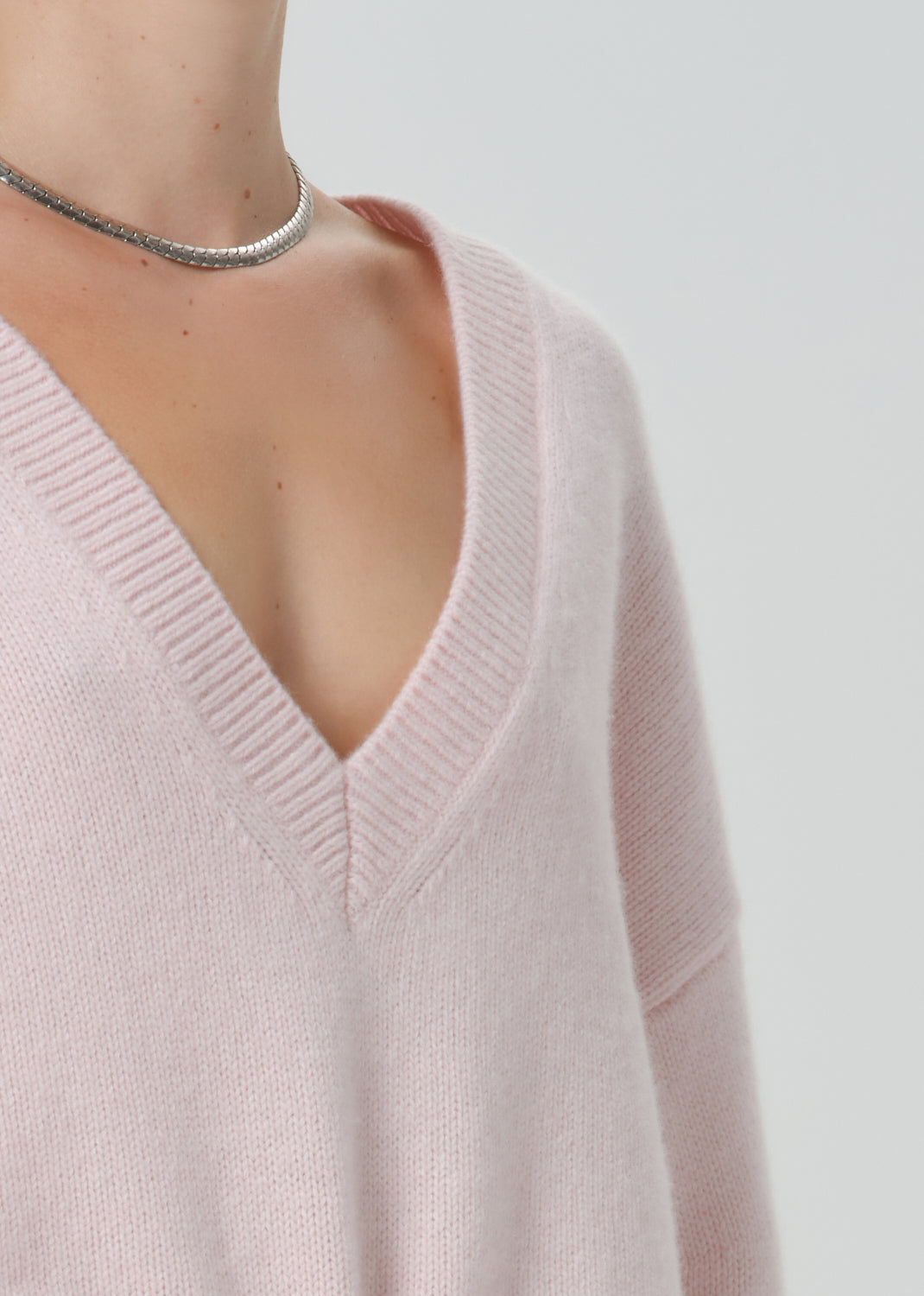 Close-up of a person wearing a pale blush pink V-neck knit sweater, with a silver chain choker visible against a light grey background.