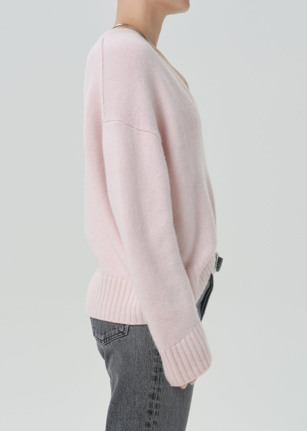 A side-profile view of a person wearing a light pink knit sweater with ribbed cuffs and hem, paired with grey jeans, against a grey background.