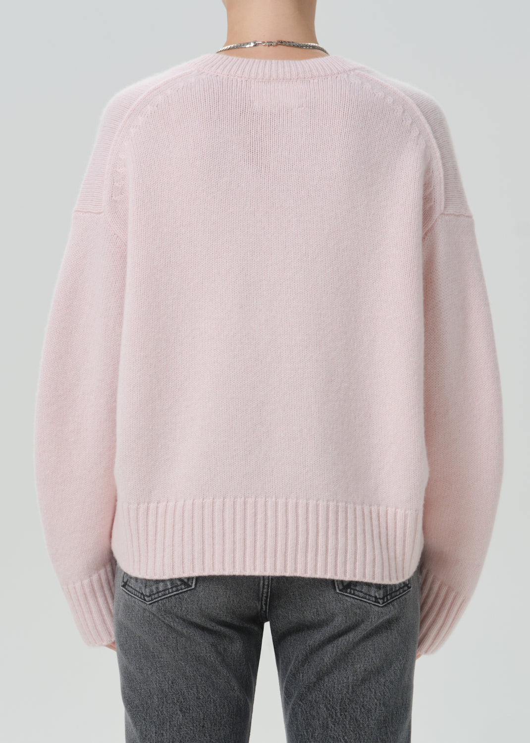 A person shown from the back wearing a light pink knit sweater and dark grey jeans against a plain light grey background.