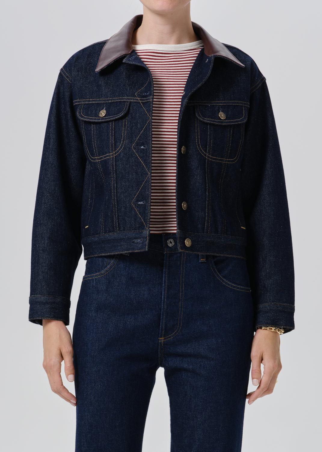 A model wearing a dark indigo denim jacket with a brown collar, layered over a red and white striped t-shirt and paired with matching dark blue jeans.