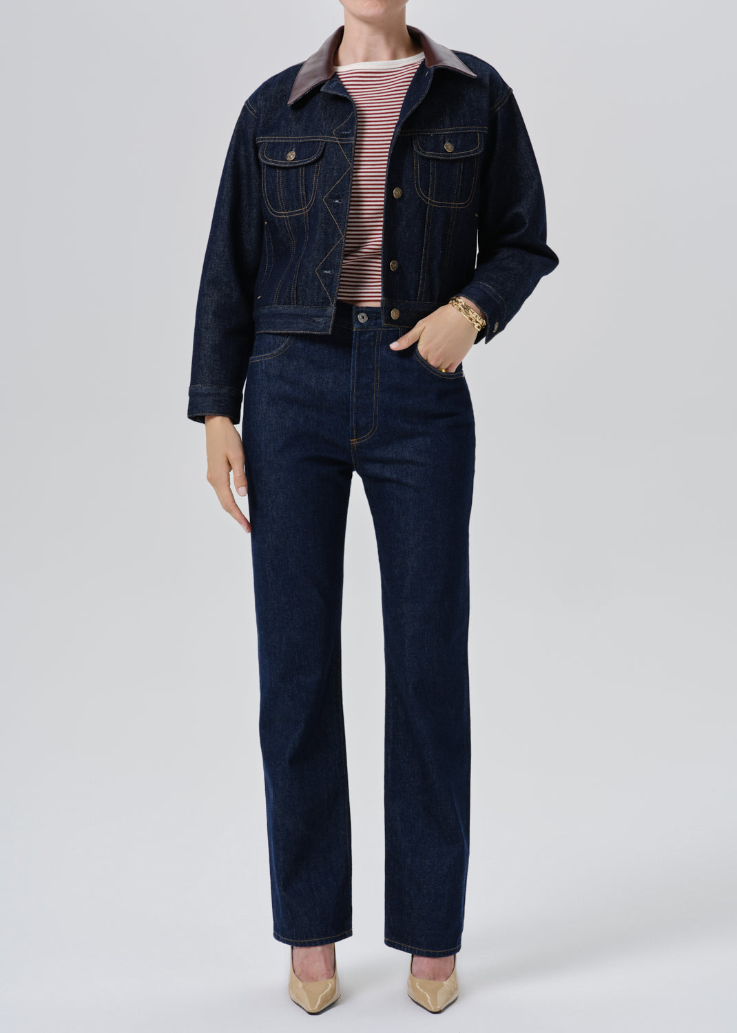 A model wearing a dark indigo denim set consisting of a cropped button-up jacket with a contrasting brown collar and matching straight-leg jeans, styled with a pink and white striped t-shirt and beige heels against a grey background.