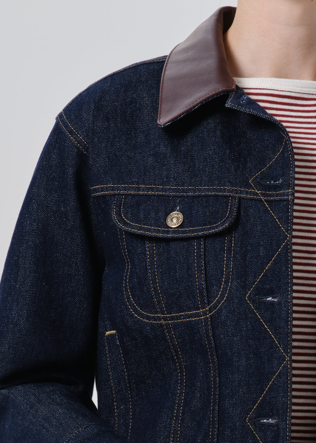 Close-up of a dark indigo denim jacket with a contrasting brown collar, featuring contrast stitching and a buttoned chest pocket, worn over a red and white striped shirt.