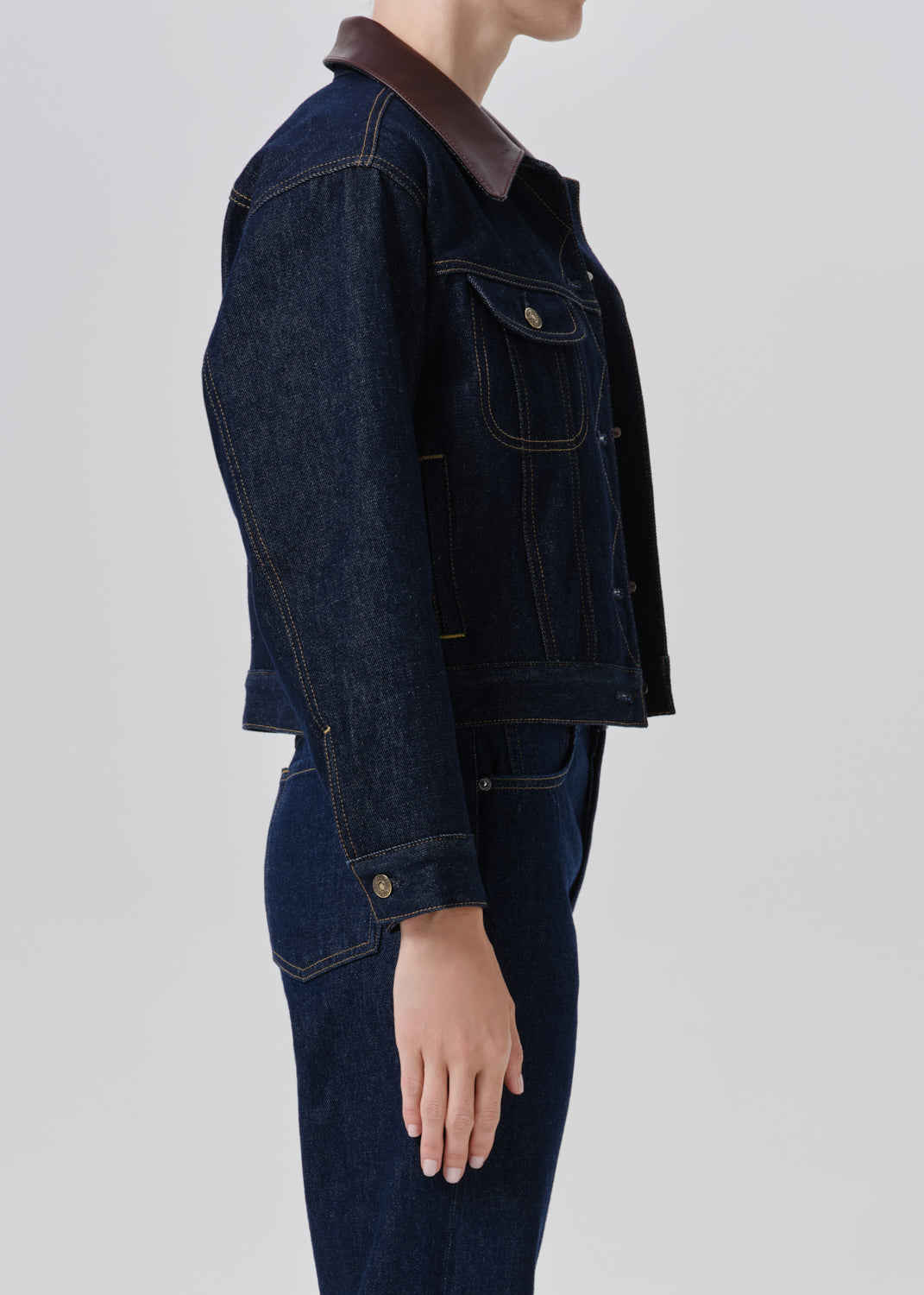 A side-profile view of a model wearing a dark indigo denim jacket with a contrasting brown collar, paired with matching dark denim jeans, against a grey background.