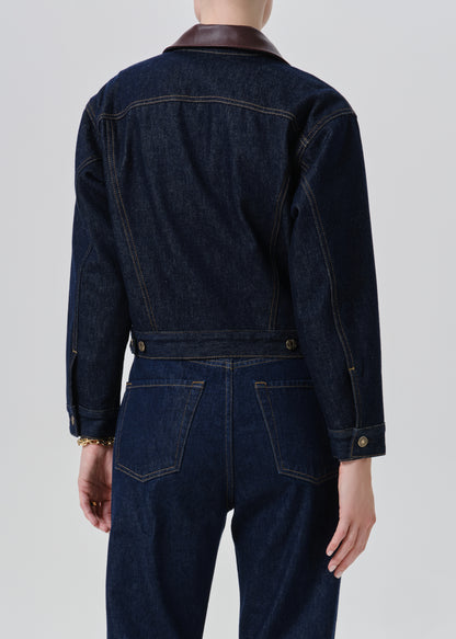Back view of a person wearing a dark indigo denim jacket with a brown collar and matching dark denim pants, photographed against a grey background.