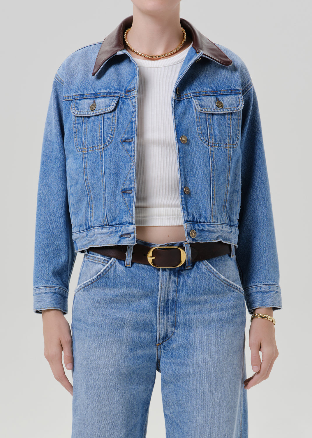 Person wearing a light wash denim jacket with a white top and matching light wash denim jeans on a grey background