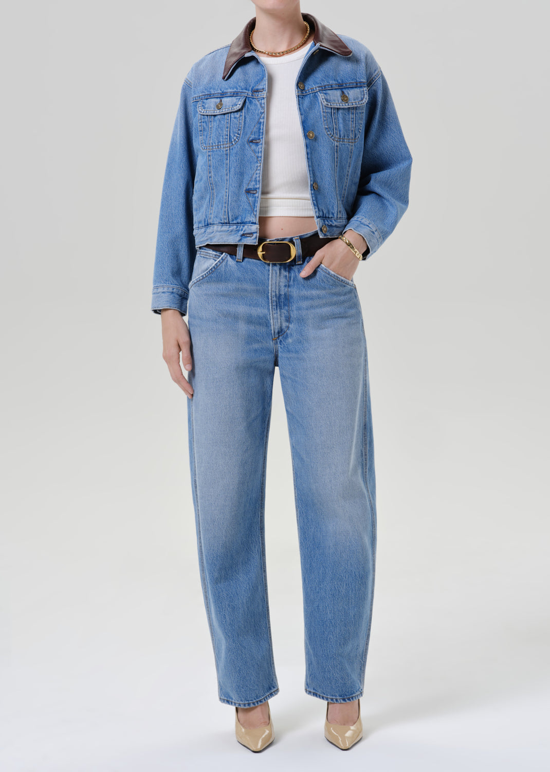 Person wearing a light wash denim jacket with a white top matching light wash denim jeans and beige pointed toe heels on a grey background