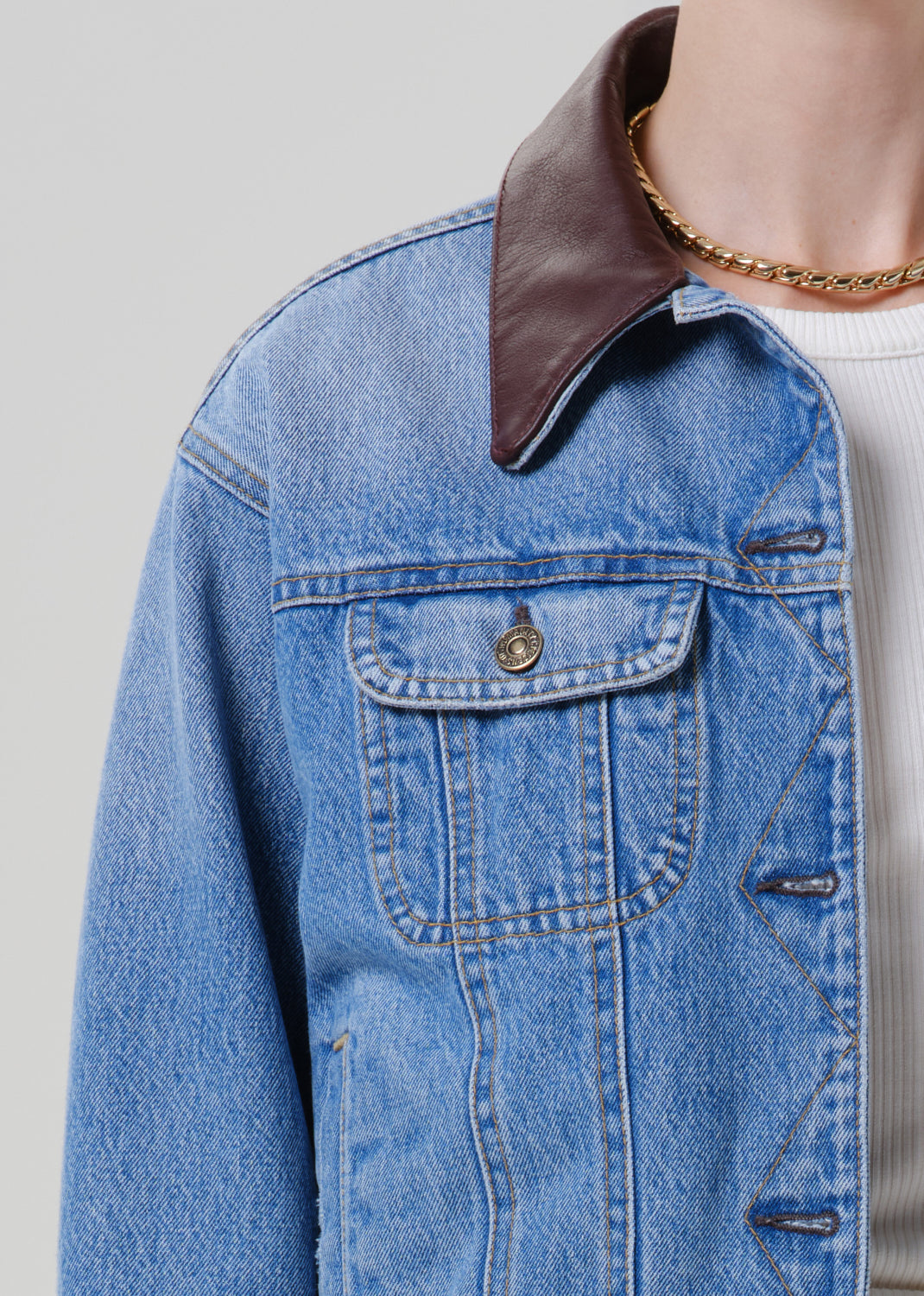 Person wearing a light wash denim jacket with a brown leather collar on a grey background