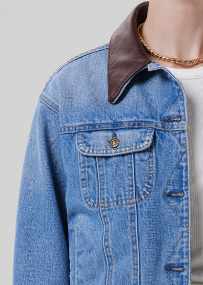 Person wearing a light wash denim jacket with a brown leather collar on a grey background