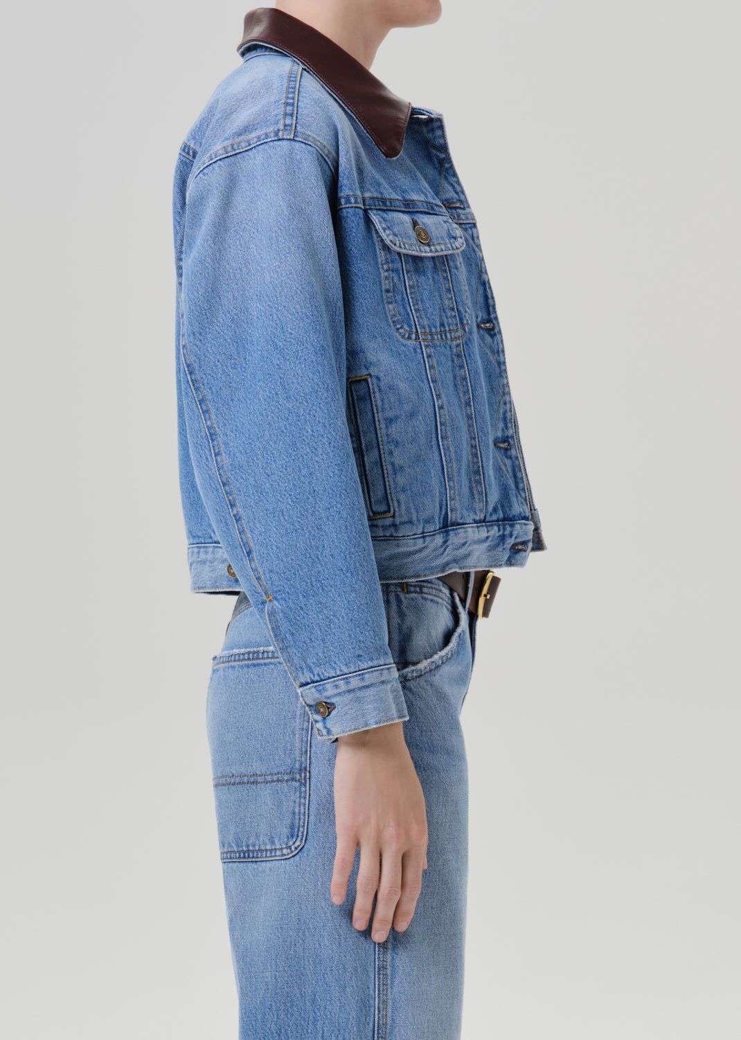 Person wearing a light wash denim jacket with matching light wash denim jeans viewed from the side on a grey background