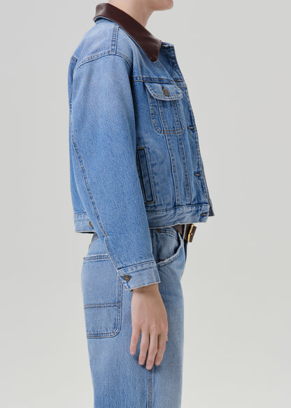 Person wearing a light wash denim jacket with matching light wash denim jeans viewed from the side on a grey background