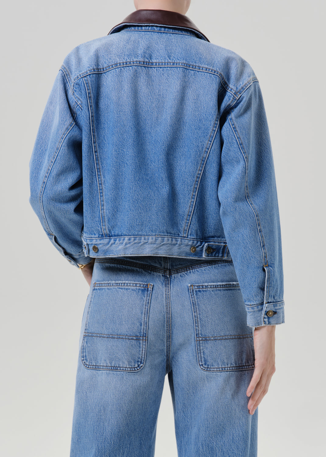 Person wearing a light wash denim jacket with matching light wash denim jeans viewed from the back on a grey background