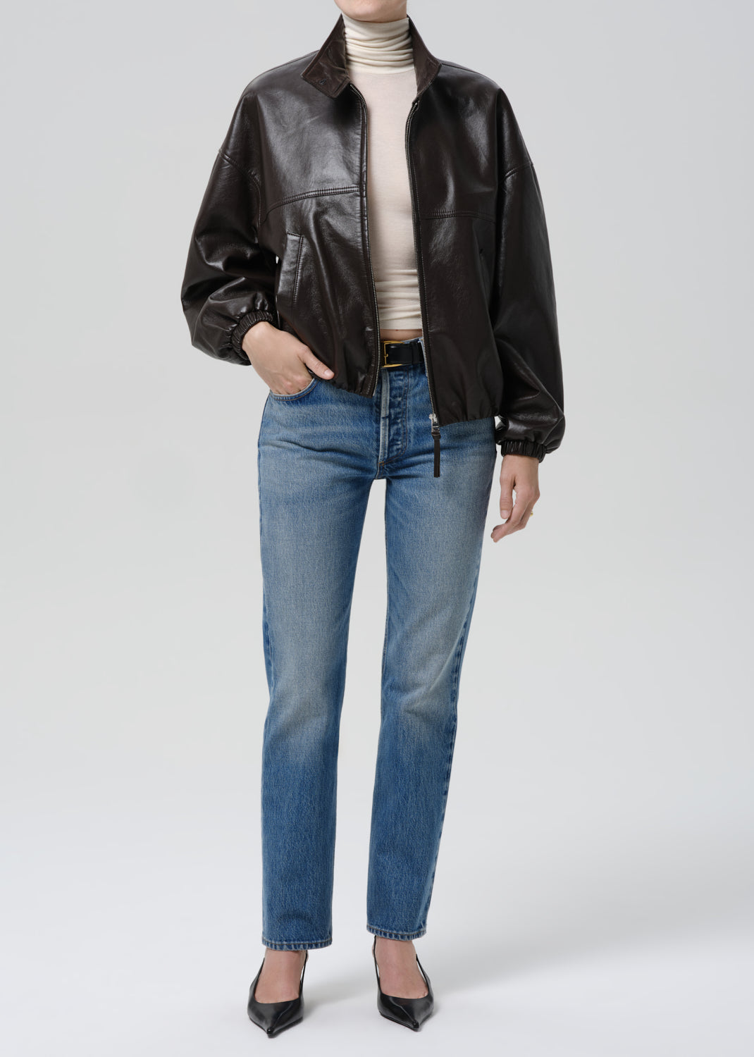 Person wearing a brown leather jacket, beige turtleneck, blue jeans, and black shoes on a grey background