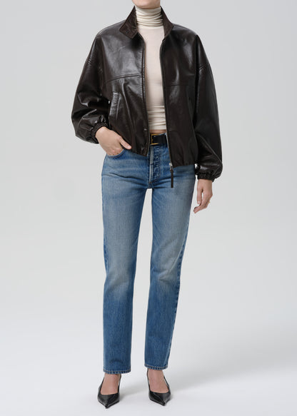 Person wearing a brown leather jacket, beige turtleneck, blue jeans, and black shoes on a grey background