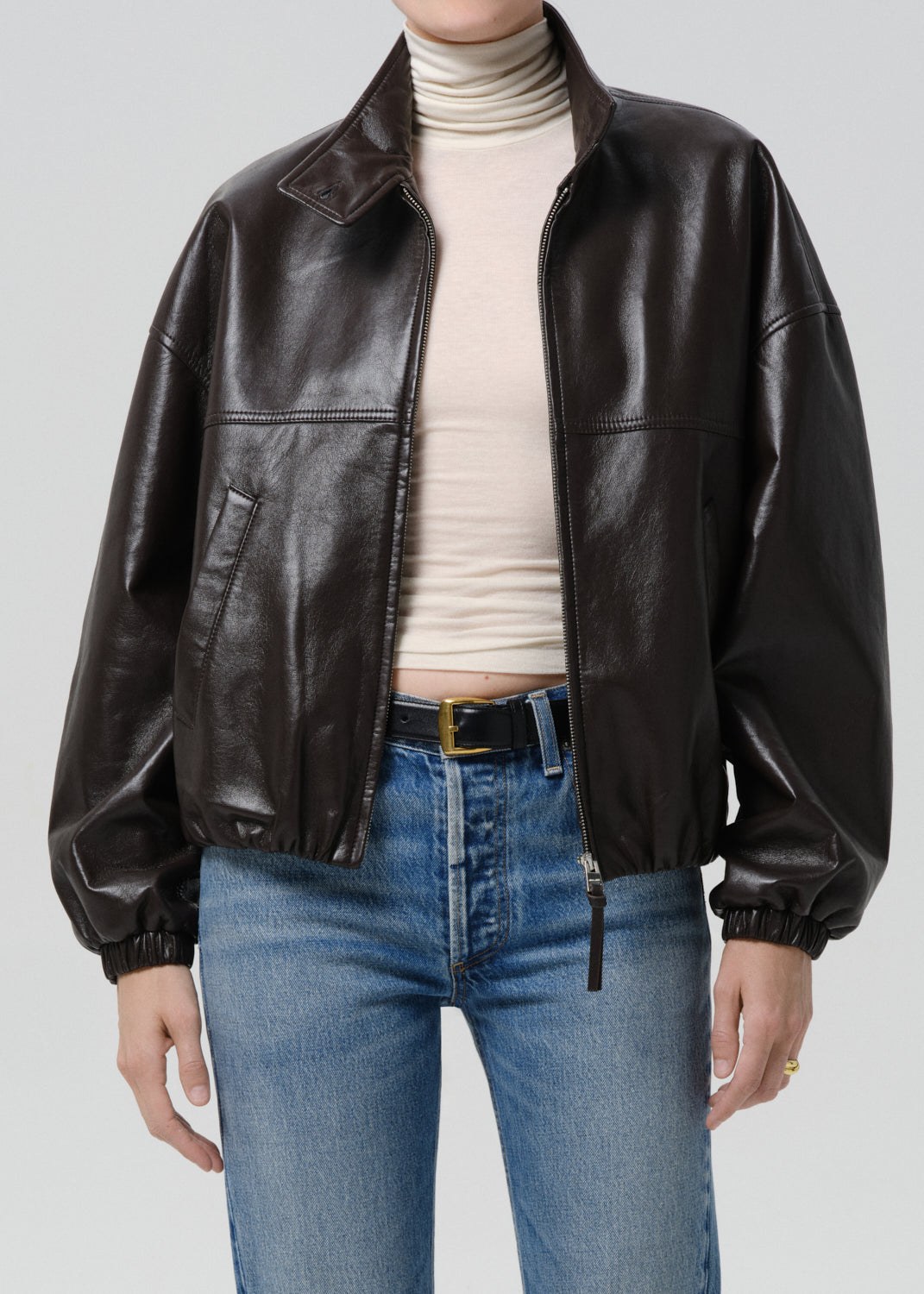Person wearing a brown leather jacket, white turtleneck, and blue jeans on a grey  background