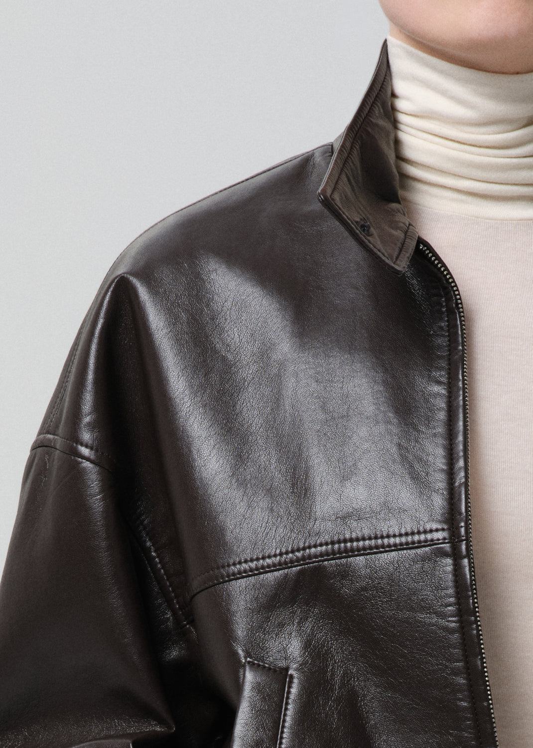 Close-up of a brown leather jacket worn over a white turtleneck on a grey background