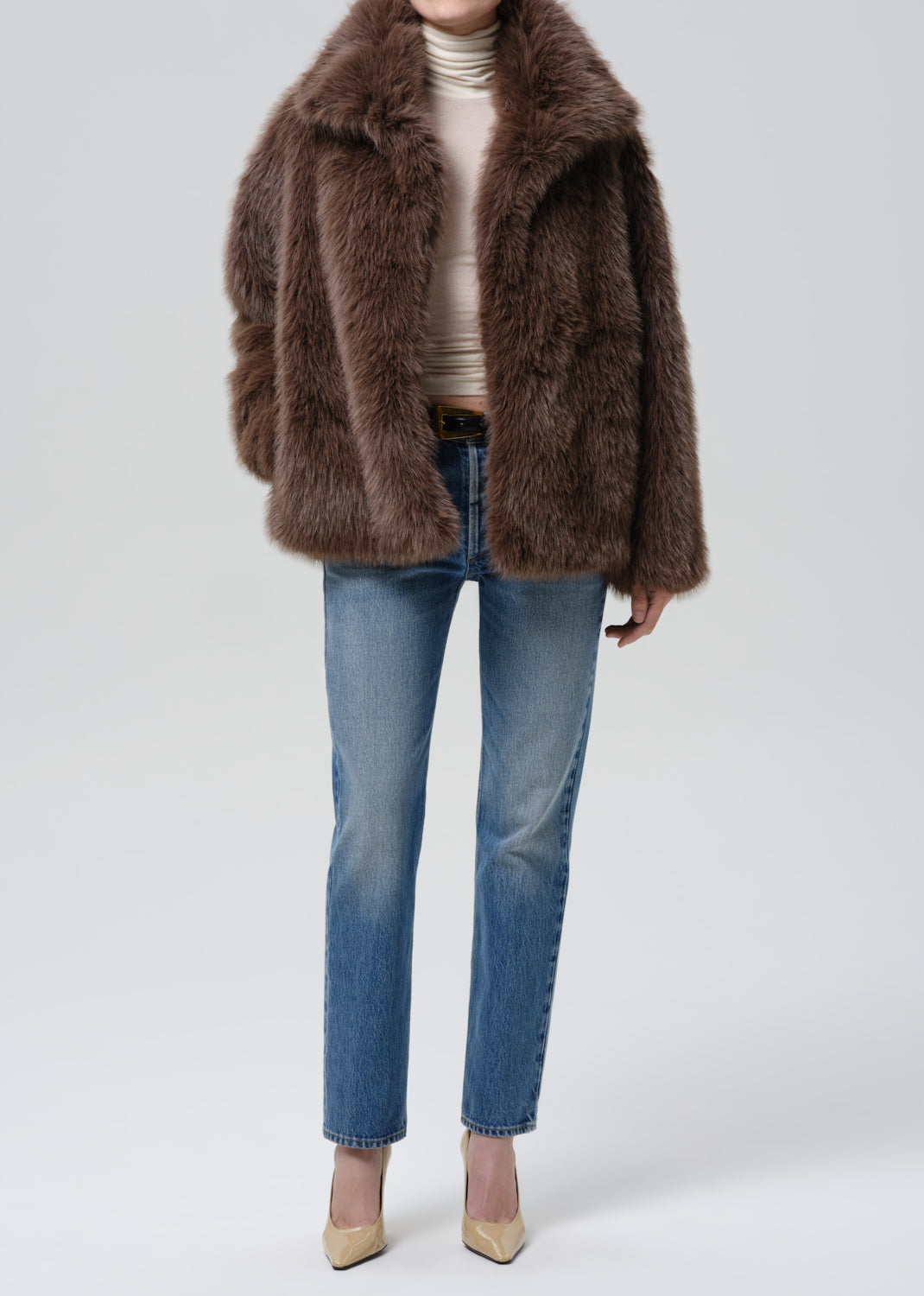 Person wearing a brown faux fur jacket with blue straight leg jeans on a grey background
