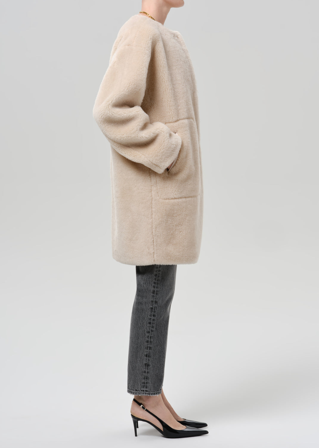 Person wearing a cream sherpa coat with grey straight leg jeans viewed from the side on a grey background