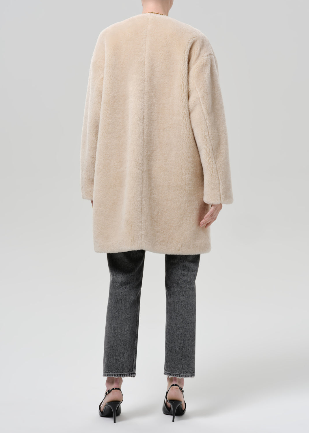 Person wearing a cream sherpa coat with grey straight leg jeans viewed from the back on a grey background