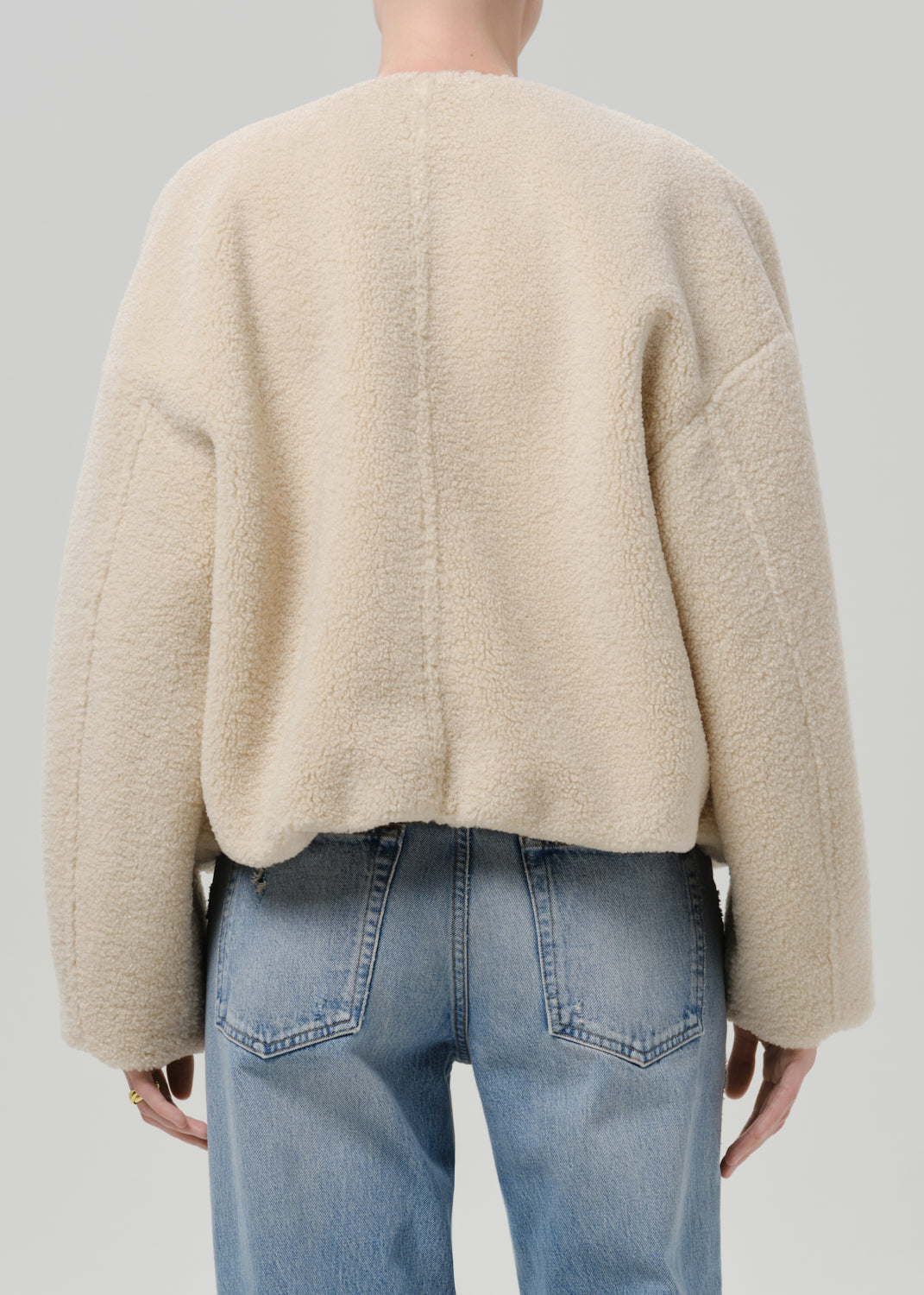 Person wearing a cream boucle jacket with light blue straight leg jeans viewed from the back on a grey background