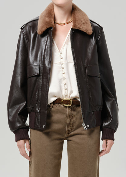 Person wearing a dark brown leather jacket with a brown shearling collar tan pants and a white blouse on a grey background