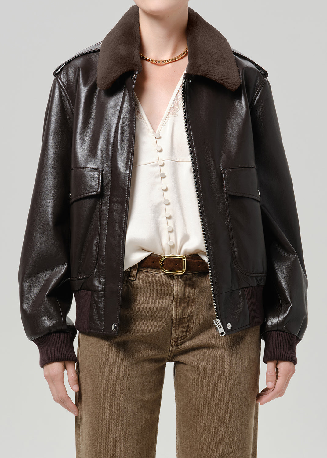 Person wearing a dark brown leather bomber jacket with a brown shearling collar, cream blouse, and brown trousers, front cropped view on a grey background.