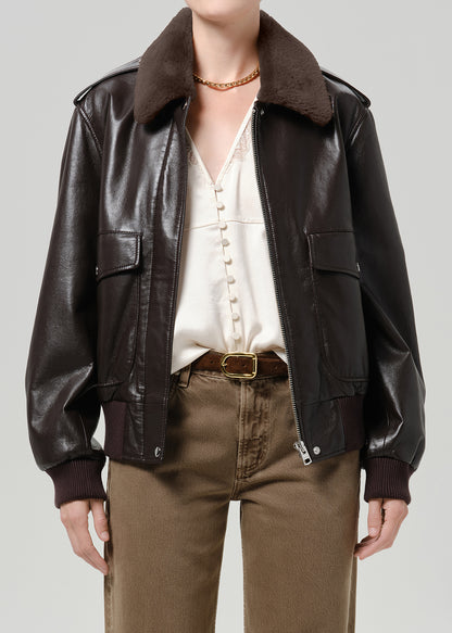 Person wearing a dark brown leather bomber jacket with a brown shearling collar, cream blouse, and brown trousers, front cropped view on a grey background.