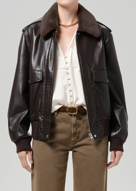 Person wearing a dark brown leather bomber jacket with a brown shearling collar, cream blouse, and brown trousers, front cropped view on a grey background.