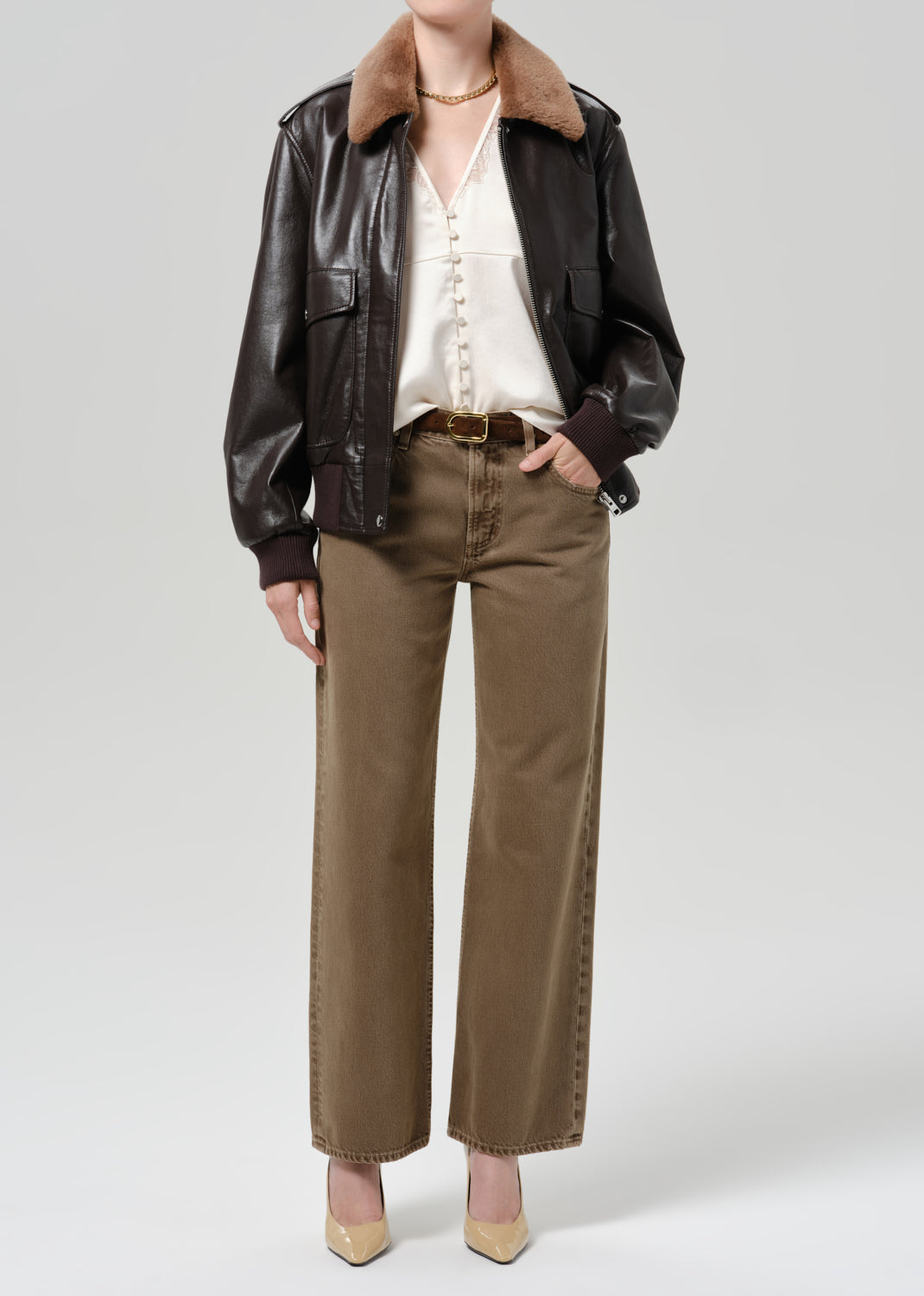 Person wearing a dark brown leather jacket with a brown shearling collar tan pants a white blouse and beige pointed toe heels on a grey background