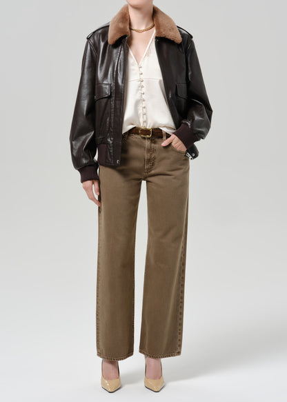 Person wearing a dark brown leather jacket with a brown shearling collar tan pants a white blouse and beige pointed toe heels on a grey background