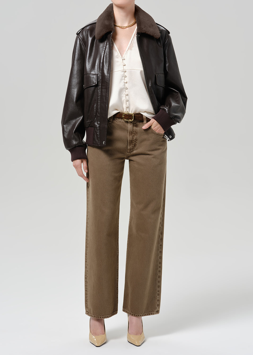 Person wearing a dark brown leather bomber jacket with a shearling collar, cream blouse, brown trousers, and light-colored shoes, front full body view on a grey background.