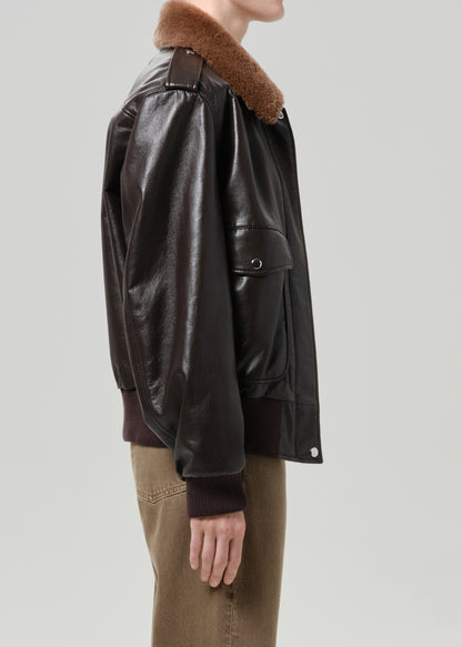Person wearing a dark brown leather jacket with a brown shearling collar and tan pants viewed from the side on a grey background
