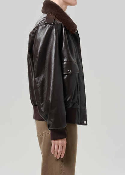 Person wearing a dark brown leather bomber jacket with a shearling collar and brown trousers, side view on a grey background.