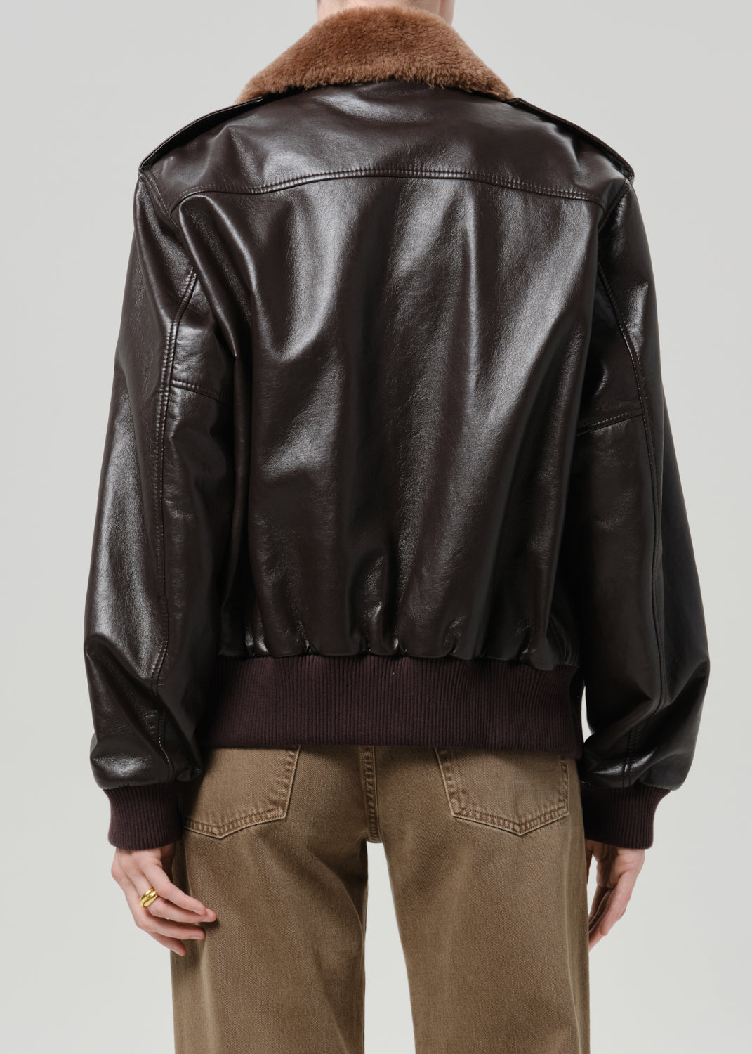 Person wearing a dark brown leather jacket with a brown shearling collar and tan pants viewed from the back on a grey background