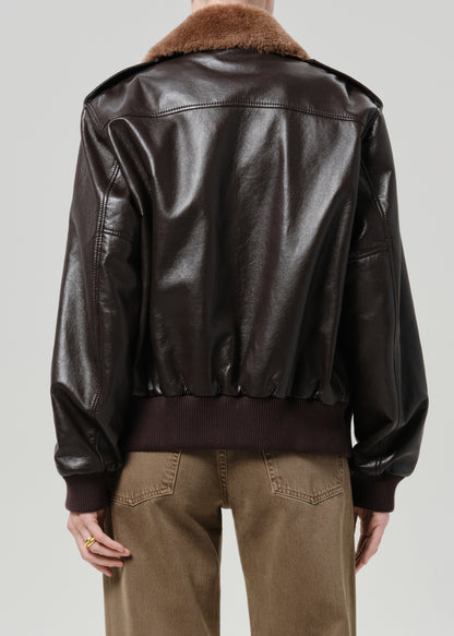 Person wearing a dark brown leather jacket with a brown shearling collar and tan pants viewed from the back on a grey background