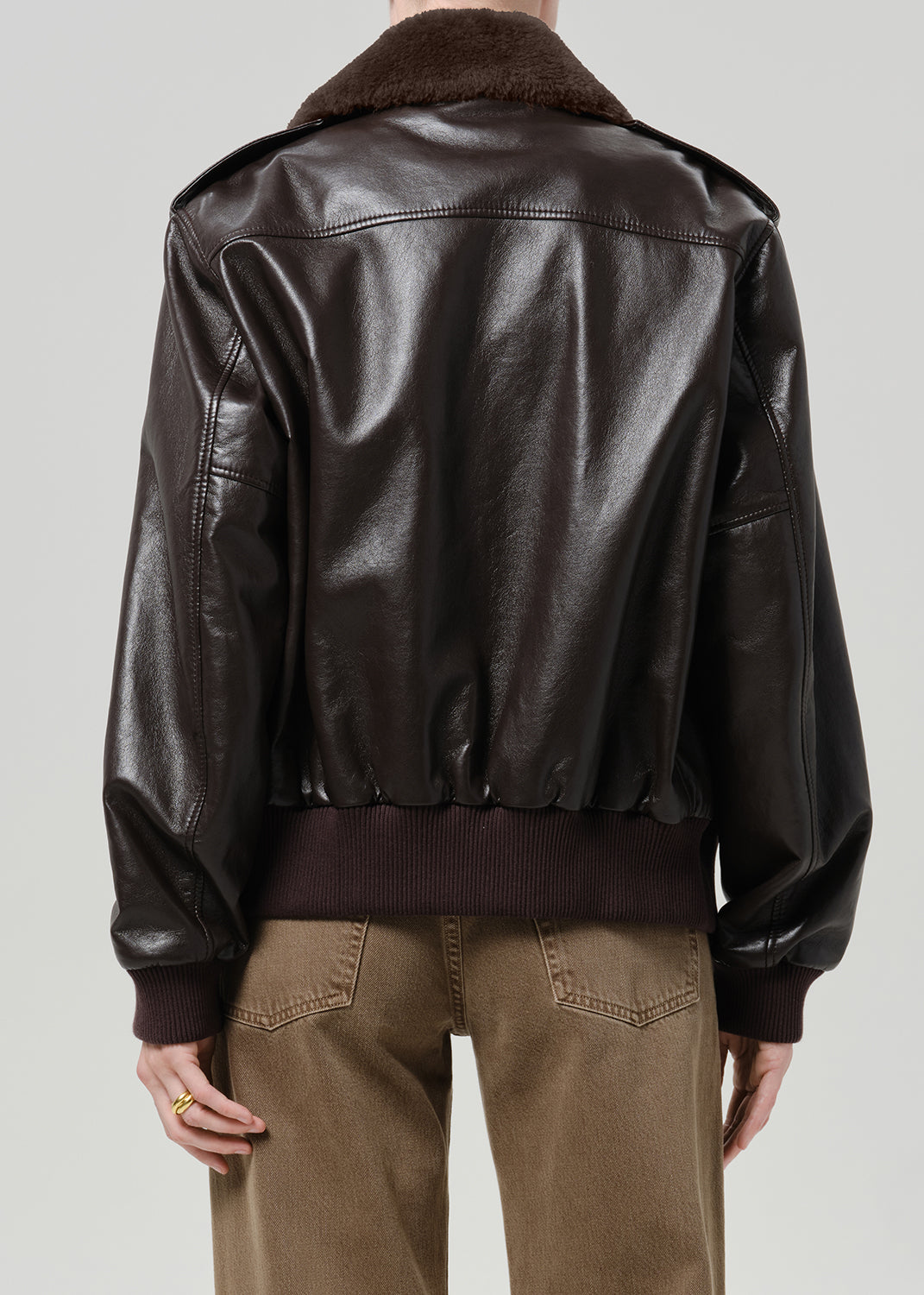 Person wearing a dark brown leather bomber jacket with a shearling collar and brown trousers, back view on a grey background.