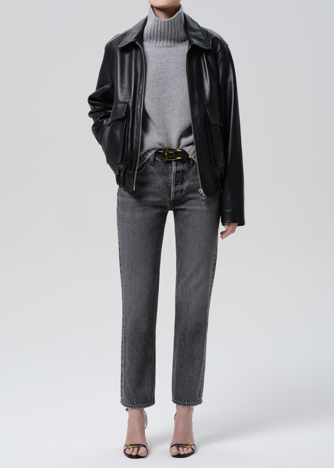 Person wearing a black leather jacket, gray turtleneck, and grey jeans on a grey background