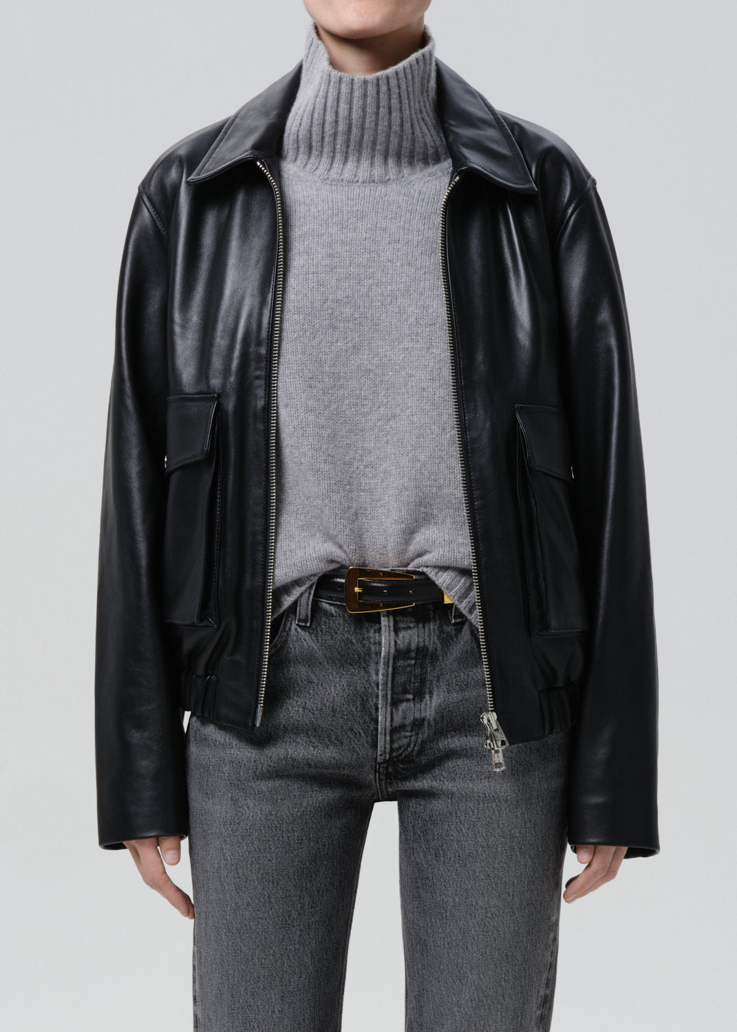 Person wearing a black leather jacket, grey sweater, and jeans on a grey background