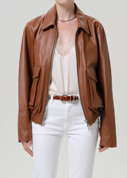 A model wearing a brown leather jacket over a cream lace-trim camisole, paired with white pants and a brown belt, against a light gray studio background.
