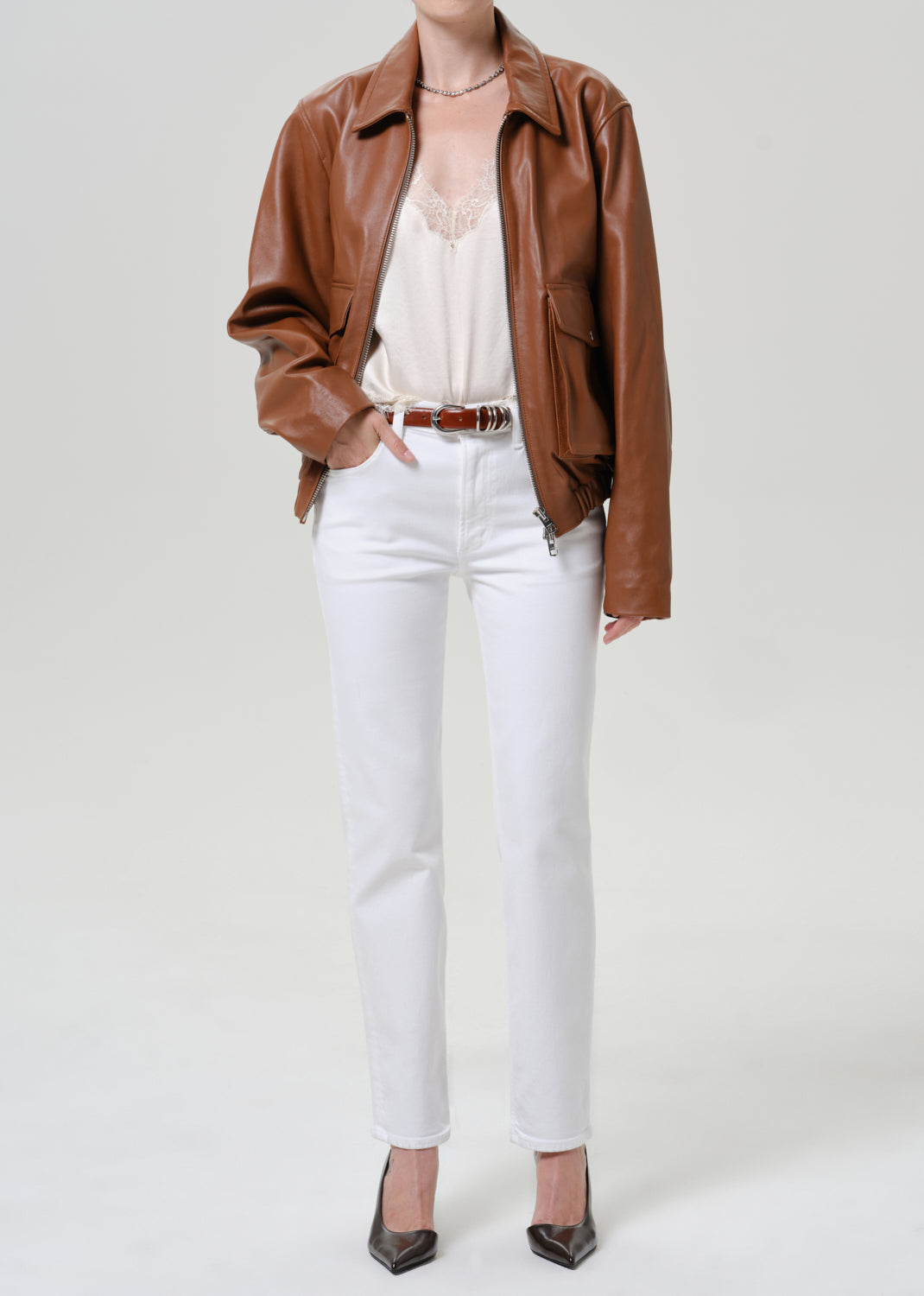 A model wearing a brown faux-leather jacket, white pants, and a cream camisole top, standing against a light gray studio background.