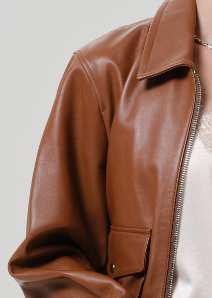 Close-up of a person wearing a smooth brown faux-leather jacket with a collar, front zipper, and flap pocket, paired with a white top against a light gray background.